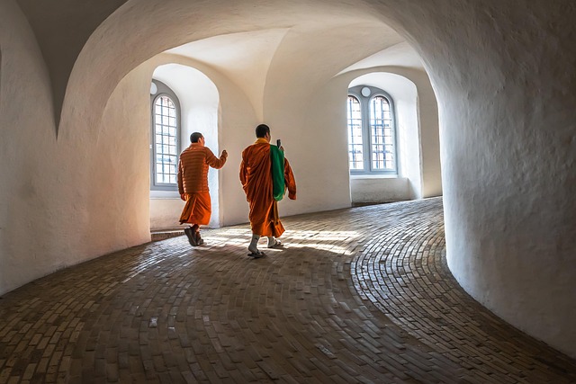 Two people in orange robes walking through a curved, arched hallway with natural light from large windows.